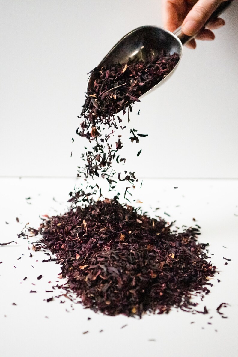 Pu&ograve; includere: Un mestolo di metallo versa foglie di t&egrave; di ibisco essiccate sfuse su una superficie bianca. Le foglie di t&egrave; sono di un rosso intenso.