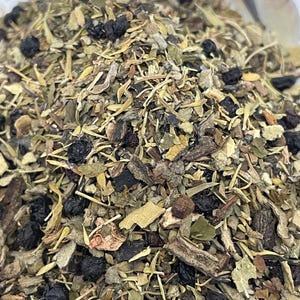 May include: A close-up of a blend of dried herbs and berries, including green leaves, brown pieces, and dark purple berries. The mixture appears to be a herbal tea blend.