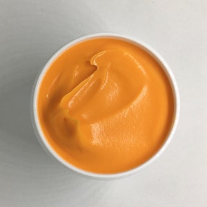 May include: A white bowl filled with a smooth, orange-colored substance. The substance has a swirl pattern and appears to be a type of cream or lotion.