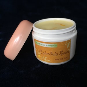 May include: A white jar with a pink lid filled with a yellow salve. The jar has a label that reads "Forest Hills Remedies Calendula Salve Net Wt. 2 oz."