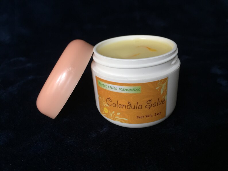 May include: A white jar with a pink lid filled with a yellow salve. The jar has a label that reads "Forest Hills Remedies Calendula Salve Net Wt. 2 oz."