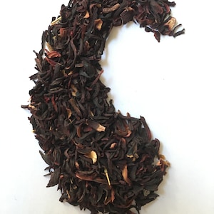 Pu&ograve; includere: Primo piano di un mucchio di fiori di ibisco secchi e altre erbe, disposti a forma di mezzaluna su uno sfondo bianco. I fiori sono di un rosso intenso e le erbe sono un mix di marrone e verde.