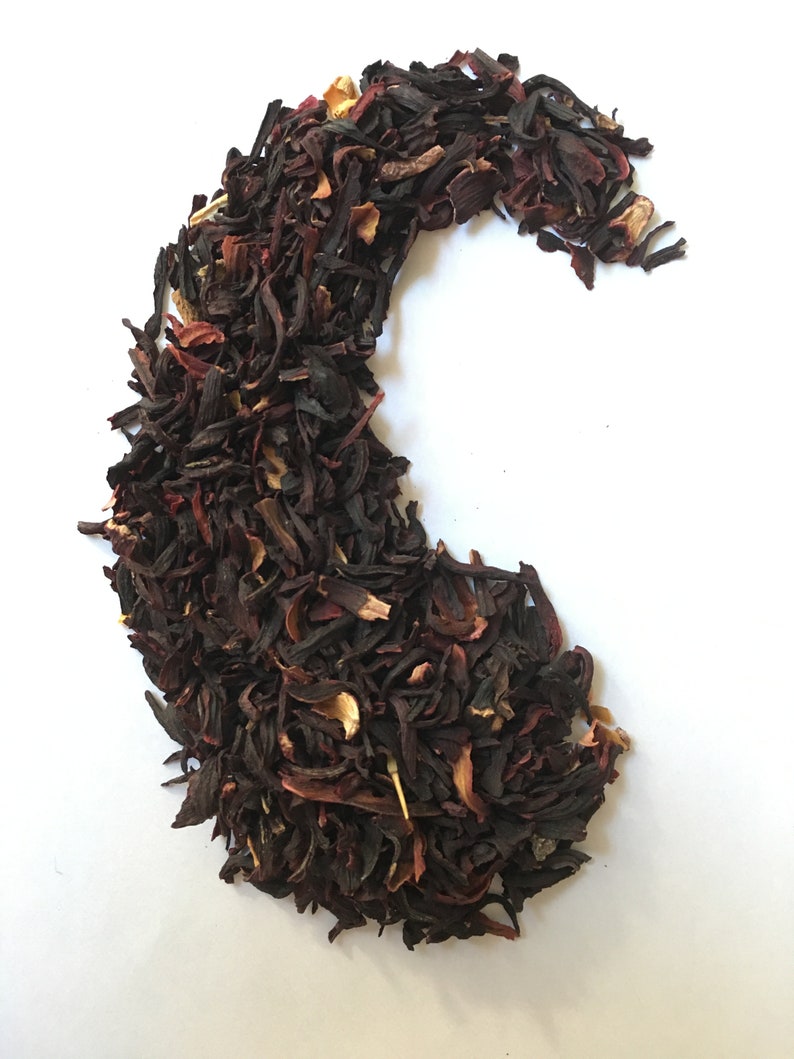 Pu&ograve; includere: Primo piano di un mucchio di fiori di ibisco secchi e altre erbe, disposti a forma di mezzaluna su uno sfondo bianco. I fiori sono di un rosso intenso e le erbe sono un mix di marrone e verde.