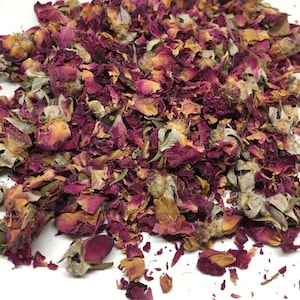 May include: Dried rose petals and buds in shades of pink, red, and brown. The petals are loose and scattered on a white surface.