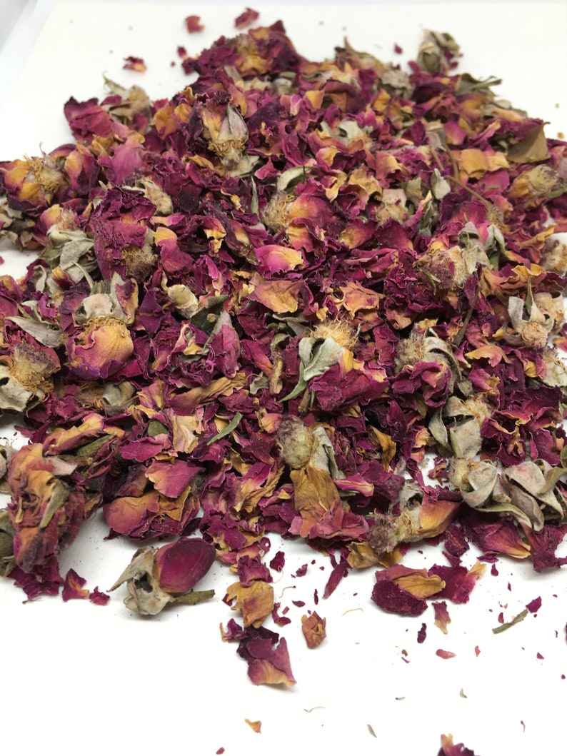 May include: Dried rose petals and buds in shades of pink, red, and brown. The petals are loose and scattered on a white surface.