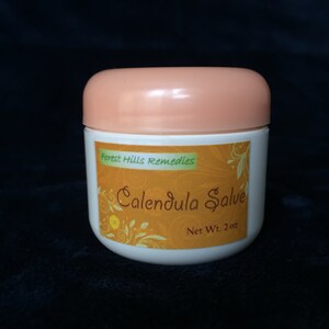 May include: A white jar with a peach-coloured lid, labelled "Forest Hills Remedies Calendula Salve Net Wt. 2 oz."