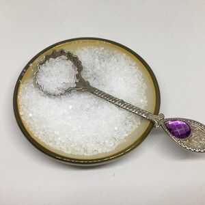 May include: A small, white ceramic dish filled with white salt crystals. A silver spoon with a purple gem on the handle rests in the dish.