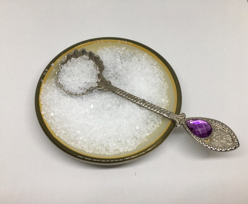 May include: A small, white ceramic dish filled with white salt crystals. A silver spoon with a purple gem on the handle rests in the dish.
