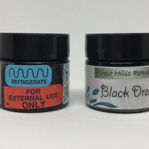 May include: Two small black jars with white labels. The jar on the left has a blue label that says "Refrigerate" and a red label that says "For External Use Only". The jar on the right has a white label with green lettering that says "Forest Hills Remedies" and "Black Drawing".