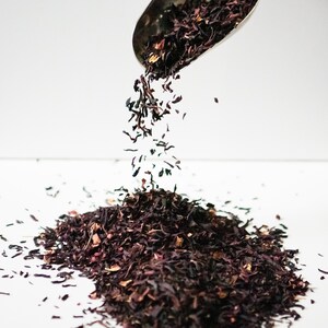 Pu&ograve; includere: Un cucchiaio di metallo versa foglie di t&egrave; di ibisco secche sfuse su una superficie bianca. Le foglie di t&egrave; sono di colore rosso scuro e hanno un aspetto leggermente strutturato.