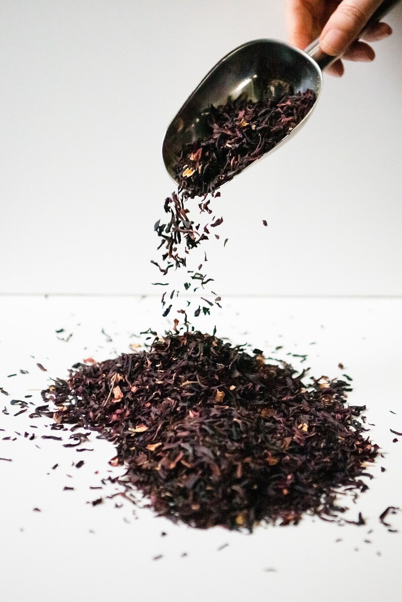 Pu&ograve; includere: Un cucchiaio di metallo versa foglie di t&egrave; di ibisco secche sfuse su una superficie bianca. Le foglie di t&egrave; sono di colore rosso scuro e hanno un aspetto leggermente strutturato.