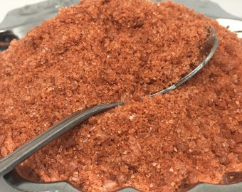 May include: Close-up of a pile of orange-red Himalayan salt crystals. The salt is in a bowl and a silver spoon is partially submerged in the salt.