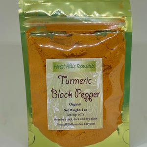 Op de afbeelding: Een verzegelde zak met groene rand gevuld met fel oranje kurkuma en zwarte peperpoeder. Het etiket luidt "Turmeric Black Pepper" en "Organic". Nettogewicht is 57 g. De zak is van Forest Hills Remedies.