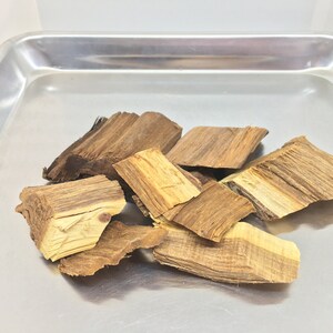 May include: A close-up of a pile of brown and light brown wood chips on a silver tray. The wood chips are irregularly shaped and have a rough texture.
