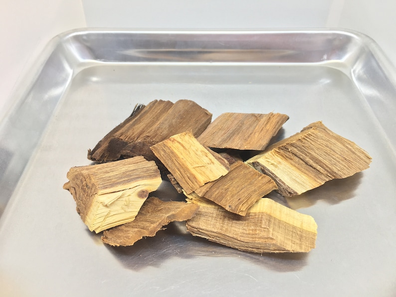 May include: A close-up of a pile of brown and light brown wood chips on a silver tray. The wood chips are irregularly shaped and have a rough texture.