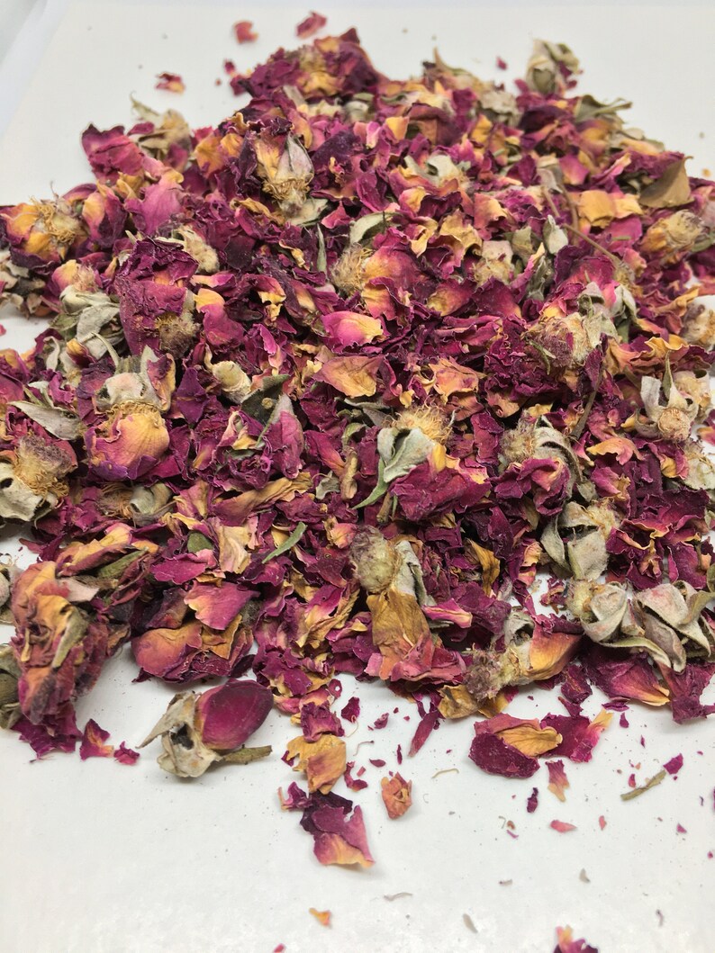 May include: A pile of dried rose petals and buds in shades of pink, red, and beige. The petals are scattered on a white surface.