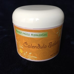 May include: A white jar with a pink lid and an orange label that says "Forest Hills Remedies Calendula Salve" with a floral design.