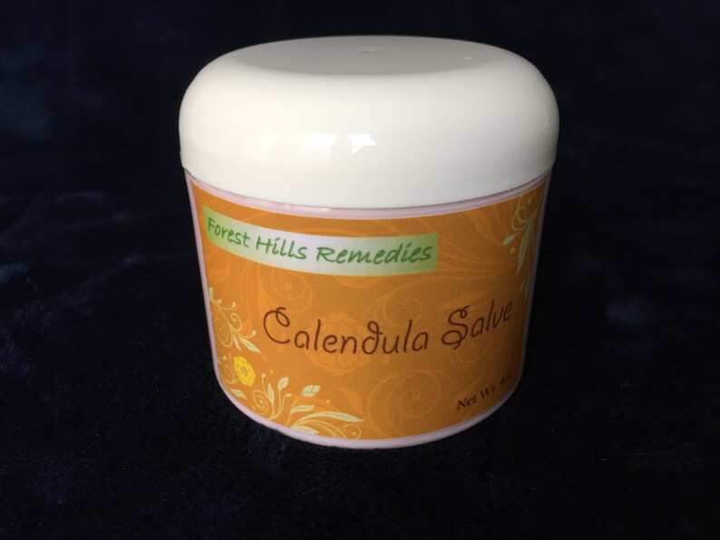 May include: A white jar with a pink lid and an orange label that says "Forest Hills Remedies Calendula Salve" with a floral design.