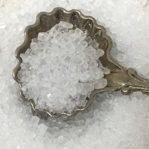 May include: A silver spoon filled with white salt crystals. The spoon is ornate with a scalloped edge and a decorative handle.