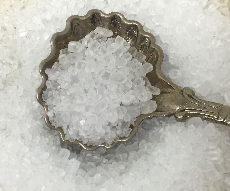 May include: A silver spoon filled with white salt crystals. The spoon is ornate with a scalloped edge and a decorative handle.
