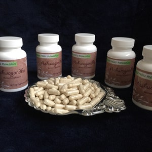 May include: Five white bottles of Ashwagandha herbal supplements from West Hills Remedies. Each bottle contains 60 vegetarian capsules. The bottles are arranged in a row on a dark blue surface. A silver dish filled with beige capsules is in the center of the bottles.