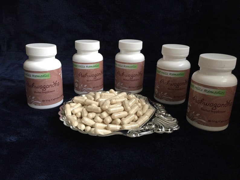 May include: Five white bottles of Ashwagandha herbal supplements from West Hills Remedies. Each bottle contains 60 vegetarian capsules. The bottles are arranged in a row on a dark blue surface. A silver dish filled with beige capsules is in the center of the bottles.