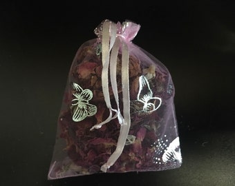 Puede incluir: Una pequeña bolsa de malla rosa llena de pétalos de rosa secos y decorada con diseños de mariposas plateadas. La bolsa está atada con una cinta blanca.
