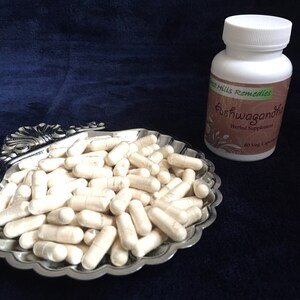 May include: A white bottle of Ashwagandha Herbal Supplement by Green Hills Remedies with 60 Veg Capsules. White capsules are displayed in a silver shell-shaped dish.