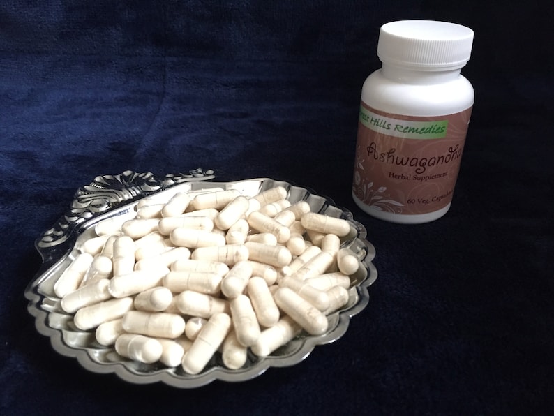 May include: A white bottle of Ashwagandha Herbal Supplement by Green Hills Remedies with 60 Veg Capsules. White capsules are displayed in a silver shell-shaped dish.