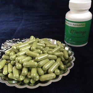 May include: A silver bowl filled with green capsules, next to a white bottle labeled "Green Hills Remedies Moringa Herbal Supplement".