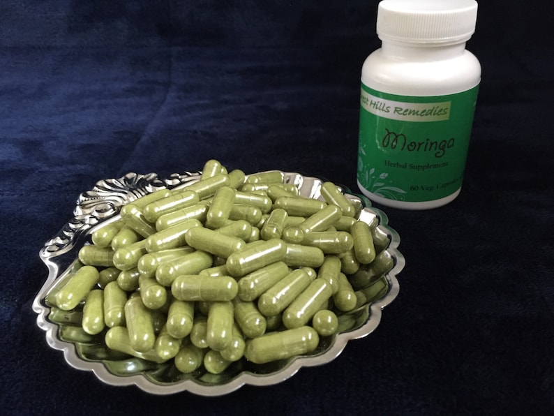 May include: A silver bowl filled with green capsules, next to a white bottle labeled "Green Hills Remedies Moringa Herbal Supplement".