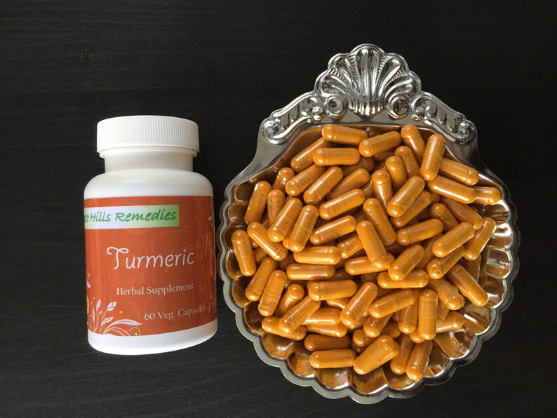 Op de afbeelding: Een witte fles met een etiket met de tekst "Hills Remedies Kurkuma Herbal Supplement 60 Veg. Capsules" en een zilveren kom gevuld met oranje kurkuma capsules.