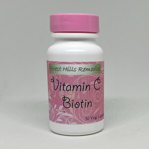 May include: A white bottle with a pink lid, labeled "Forest Hills Remedies." The label features the words "Vitamin C Biotin" in a decorative font, with a pink floral design. The bottle contains 30 capsules.