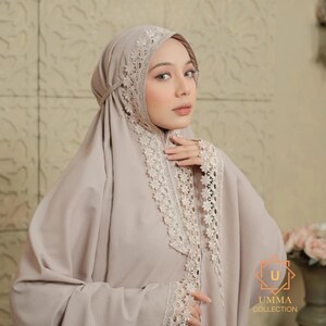 May include: A beige prayer garment and head covering with delicate lace trim. The head covering is secured under the chin. The lace trim is around the face and on the sleeves. The garment is from the Umma Collection.