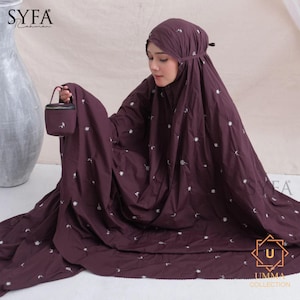 May include: A dark burgundy prayer garment with white floral embroidery. The garment includes a head covering and a matching small bag. The image features the text "SYFA Rahman" and the "UMMA COLLECTION" logo.