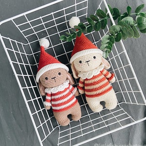 May include: Two crocheted bunny ornaments wearing Santa hats and striped sweaters. The ornaments are sitting in a white wire basket.
