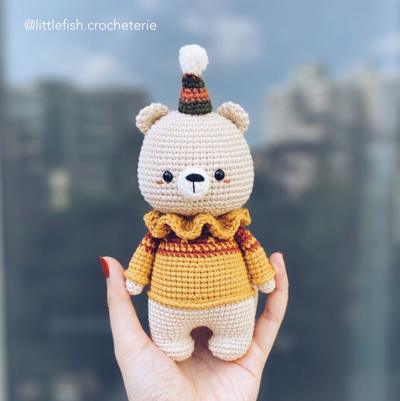 May include: A crocheted teddy bear wearing a yellow and orange striped sweater and a party hat. The bear has a white face with black eyes and a black nose.