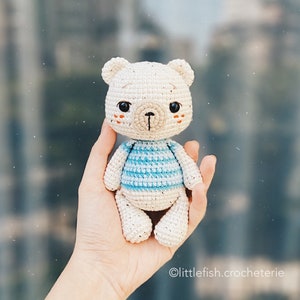 May include: A crocheted white bear wearing a blue and white striped shirt. The bear has a brown nose and black eyes.