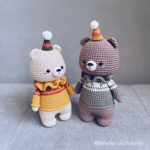 May include: Two crocheted teddy bears wearing colorful hats and sweaters. The bear on the left is white with a yellow and red sweater, while the bear on the right is brown with a green and white sweater.