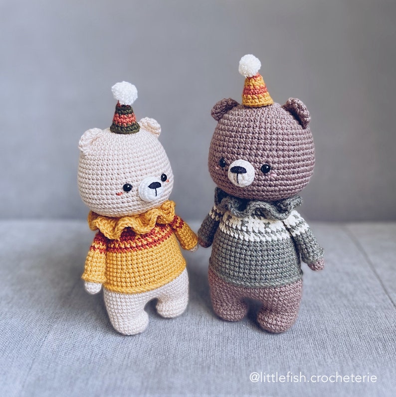 May include: Two crocheted teddy bears wearing colorful hats and sweaters. The bear on the left is white with a yellow and red sweater, while the bear on the right is brown with a green and white sweater.