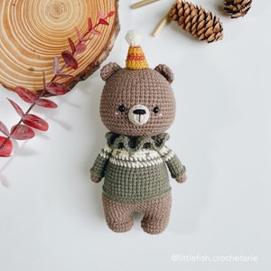 May include: A brown crocheted bear wearing a green and white sweater and a yellow and orange party hat. The bear has black eyes and a black nose.