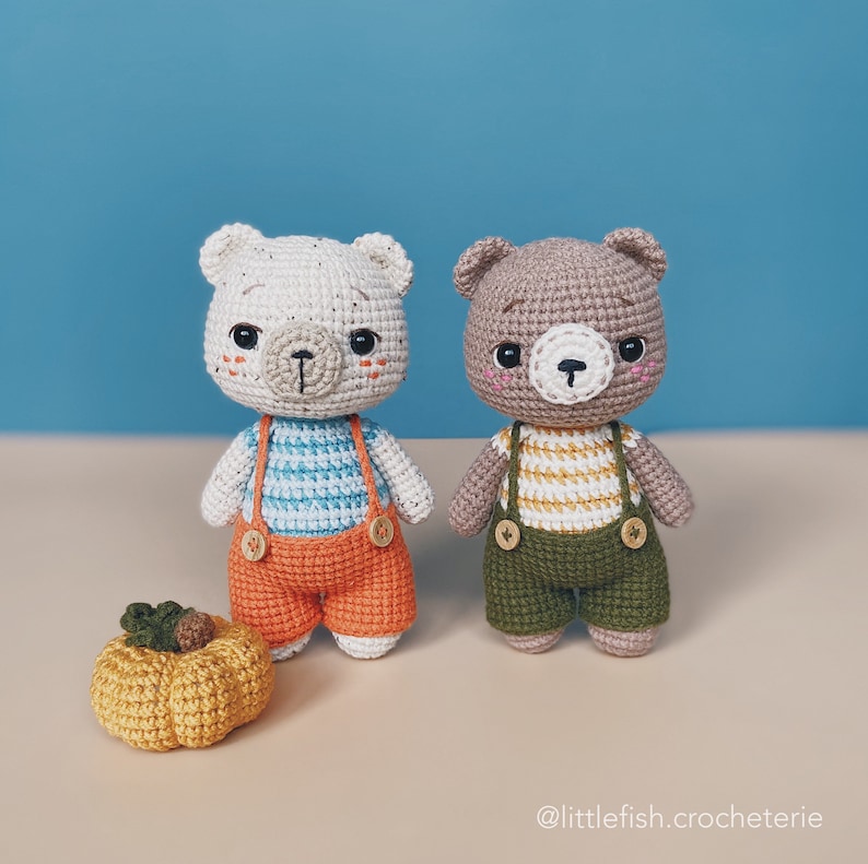 May include: Two crocheted teddy bears wearing overalls. One bear is white with blue and white striped shirt and orange overalls. The other bear is brown with a yellow and white striped shirt and green overalls. A small crocheted pumpkin sits in front of the bears.