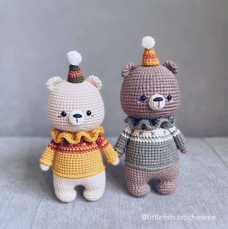 May include: Two crocheted teddy bears wearing party hats and sweaters. The bear on the left is wearing a yellow sweater with a red and orange stripe and a white pom-pom hat. The bear on the right is wearing a green sweater with a white and brown stripe and a white pom-pom hat.  @littlefish.crocheterie