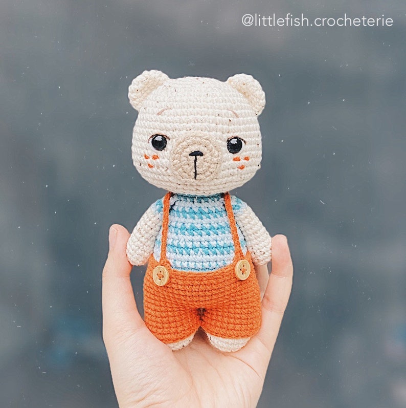 May include: A crocheted stuffed bear toy wearing orange overalls with blue and white striped shirt. The bear has a white face with brown eyes and a brown nose.