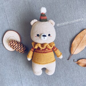May include: A crocheted teddy bear wearing a yellow sweater with a red and brown collar and a green and brown hat. The bear has a white face with black eyes and a black nose.