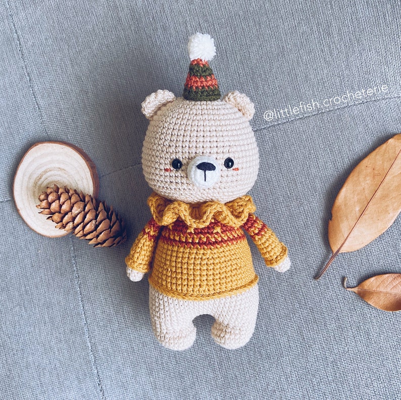 May include: A crocheted teddy bear wearing a yellow sweater with a red and brown collar and a green and brown hat. The bear has a white face with black eyes and a black nose.