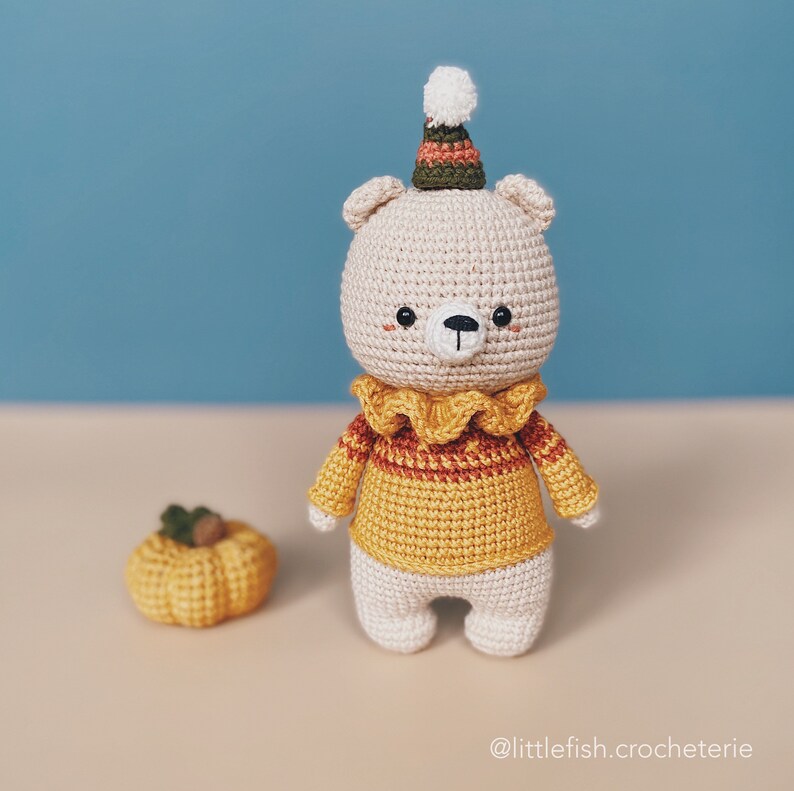 May include: A crocheted bear wearing a yellow and orange striped sweater with a white pom-pom hat. The bear is standing on a beige surface next to a small crocheted pumpkin.