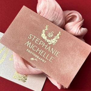 May include: Pink velvet business cards with gold foil lettering. The cards feature the name "Stephanie Michelle Photography" and a floral design. The cards are displayed on a red surface with pink fabric.