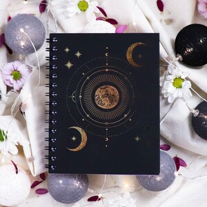 Peut inclure: Un carnet noir à spirales avec un motif céleste doré et des étoiles blanches. Le carnet est entouré de boules de coton blanches, de fleurs blanches et de fleurs roses.