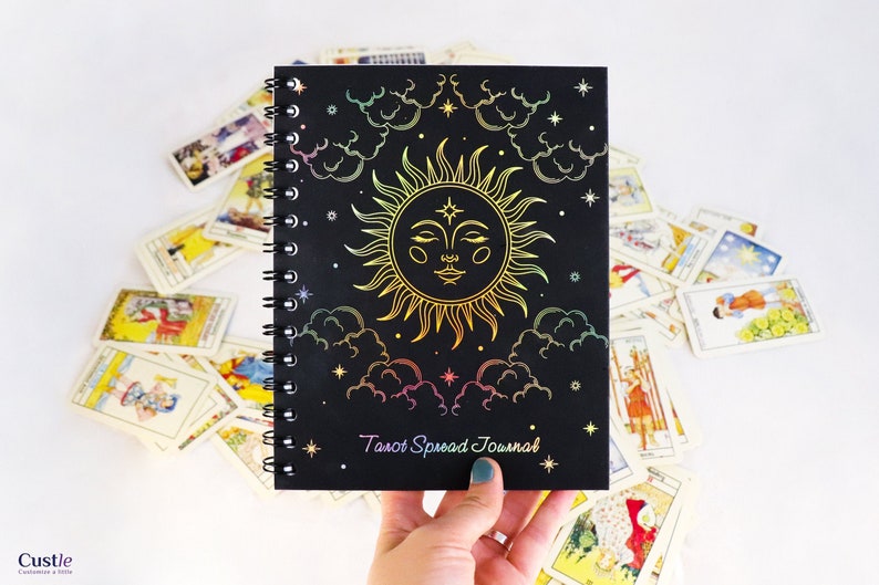 May include: A black spiral bound journal with a holographic sun and clouds design. The text "Tarot Spread Journal" is printed on the front cover.